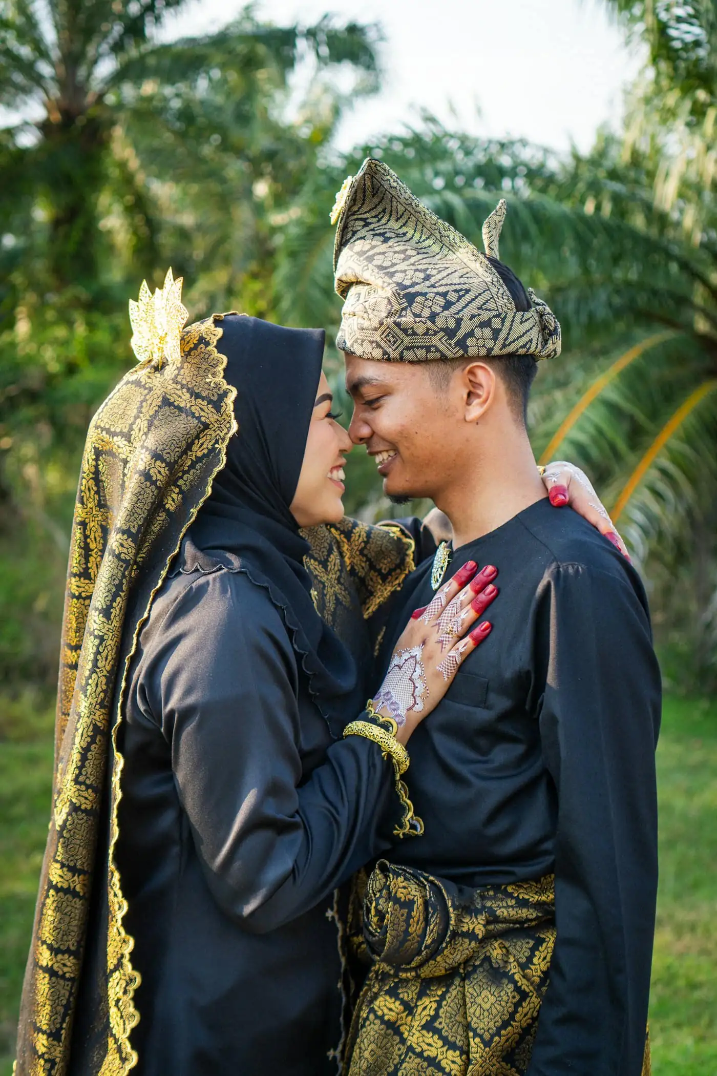 The couple in black and gold songket touching foreheads with eyes closed in a palm garden, her hand with henna on his chest