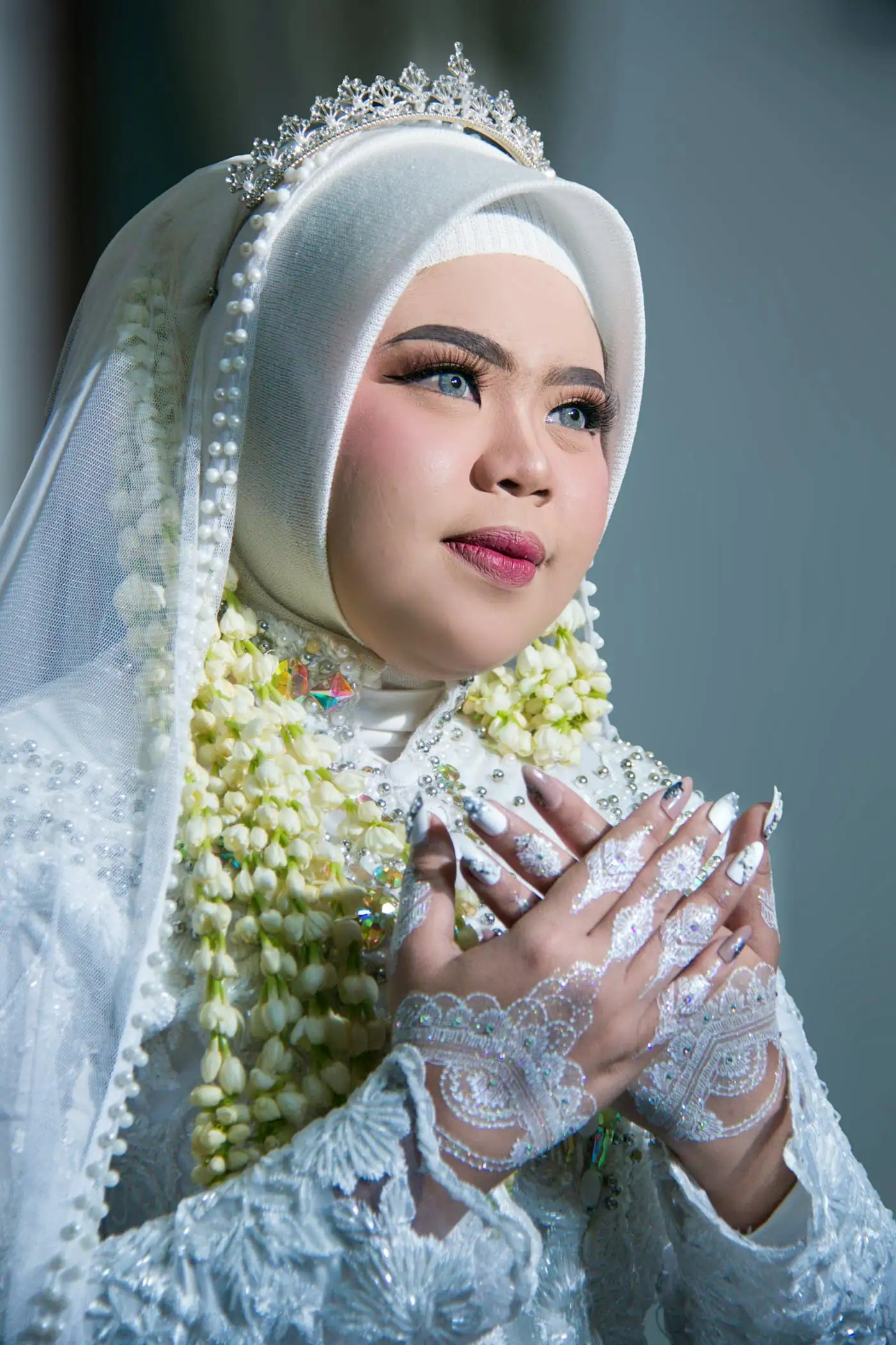 A bride in a white lace outfit and long veil with strands of jasmine, a silver crown on her hijab, crossing her lace-gloved hands over her chest