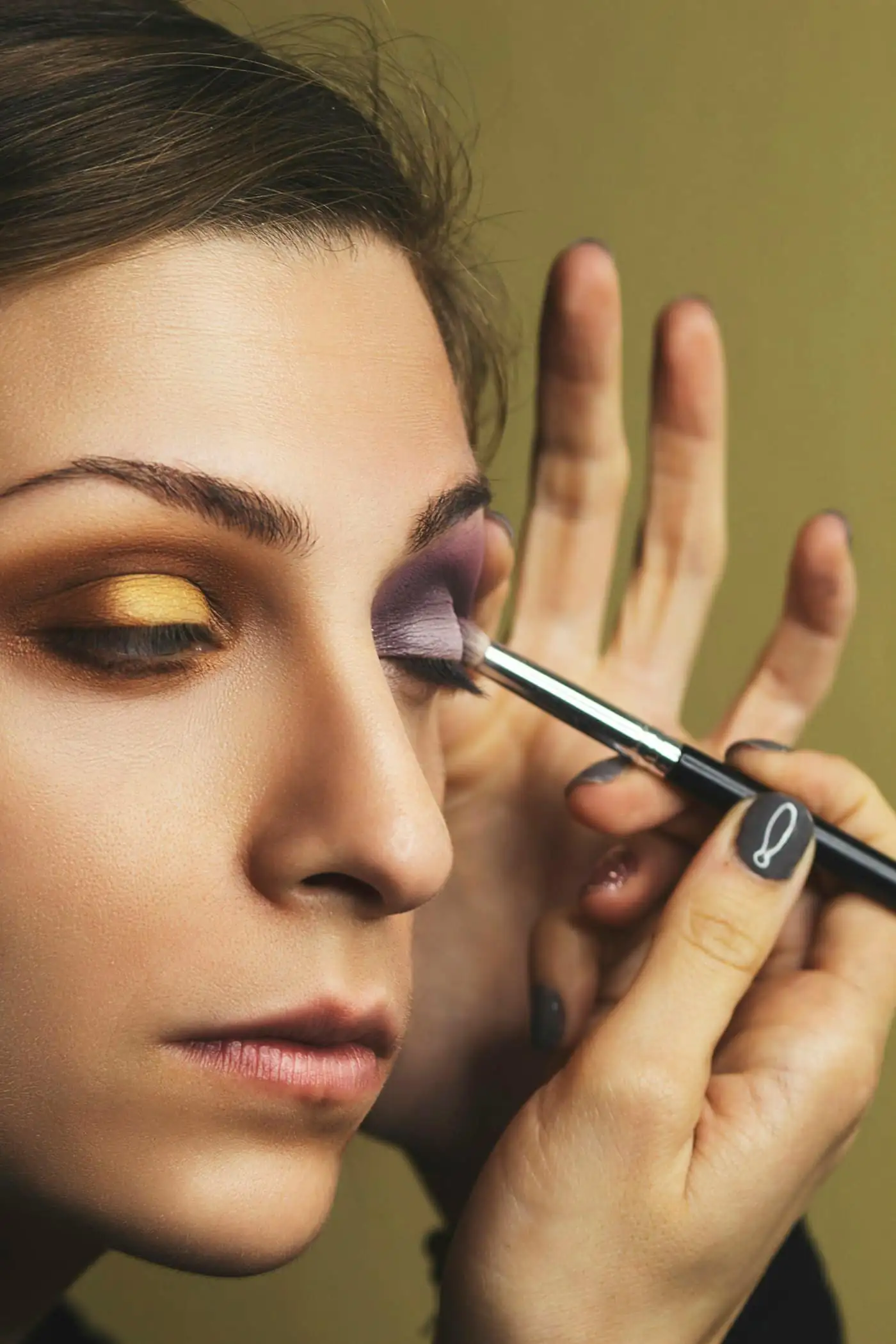 Close-up of a makeup artist’s hand blending gold and violet eyeshadow onto a woman’s eyelid with a fine brush