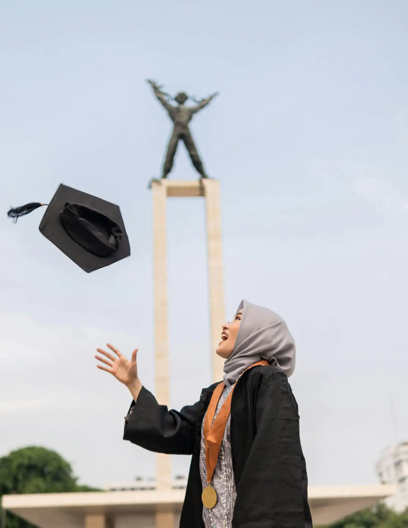 A graduate in a grey hijab and black gown with a medal throwing her mortarboard into the air beside a tall monument
