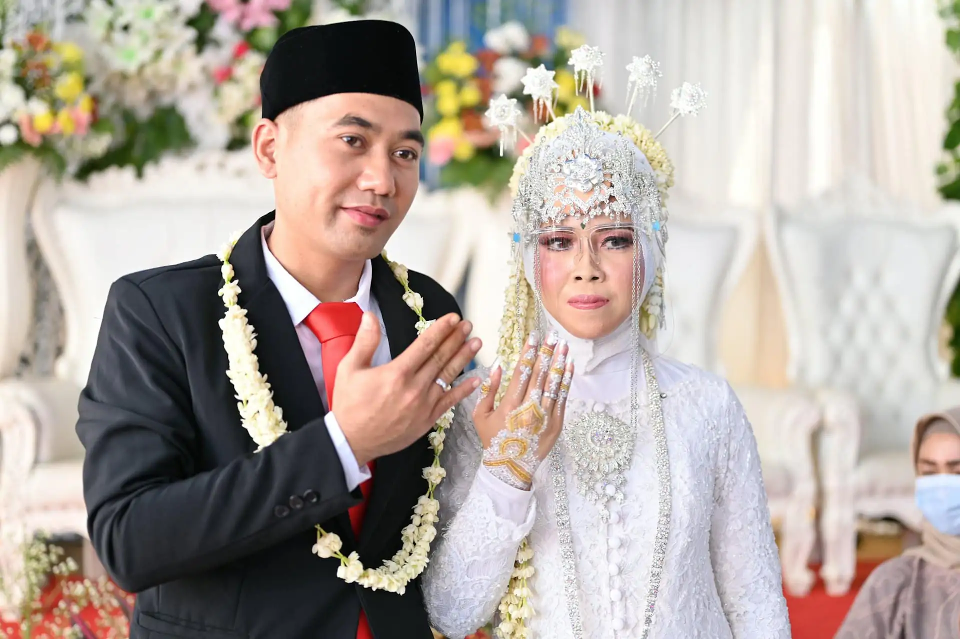 A groom in a black suit, red tie and jasmine garland and a bride in white lace with a silver headpiece showing off their rings to the camera