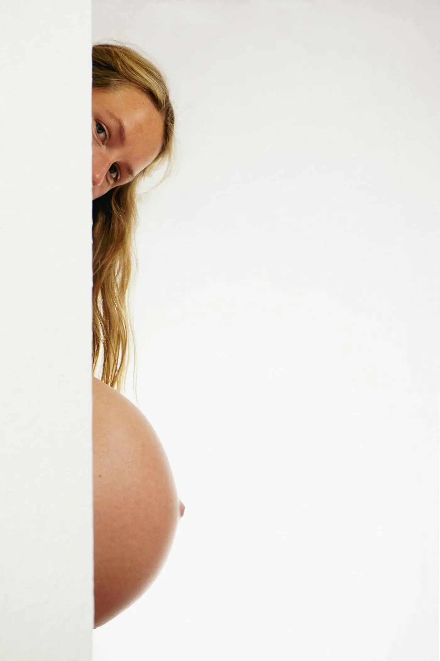 A pregnant woman with long blonde hair peeking out from behind a white wall, her bare bump in view against a plain white studio