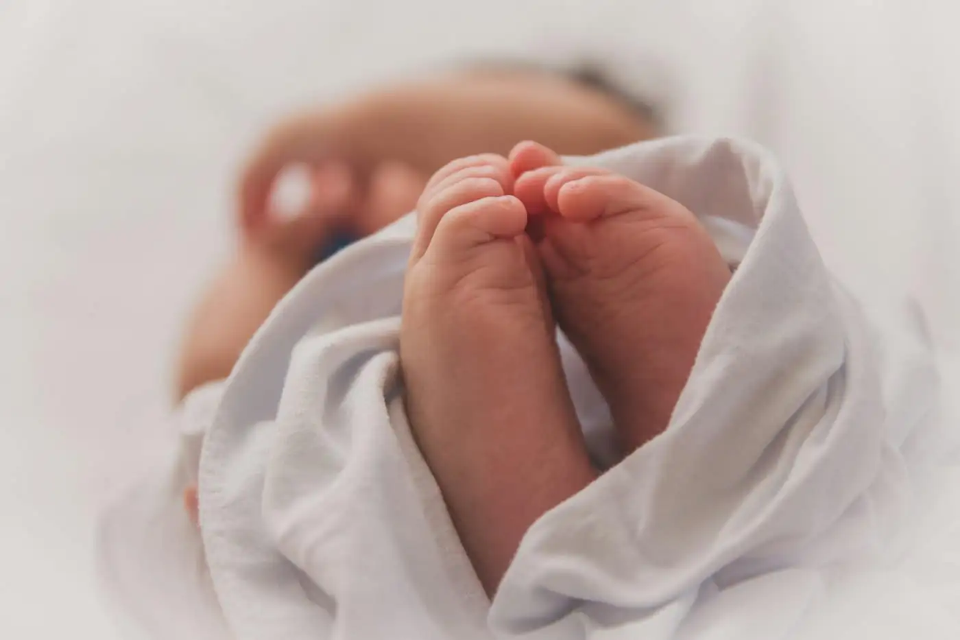 A newborn’s tiny bare feet peeking out of a white wrap, with the baby’s face softly blurred behind
