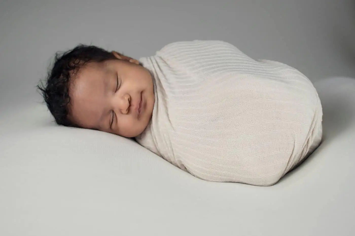 A newborn with dark hair asleep on one side, wrapped in a cream knit swaddle on a soft grey backdrop
