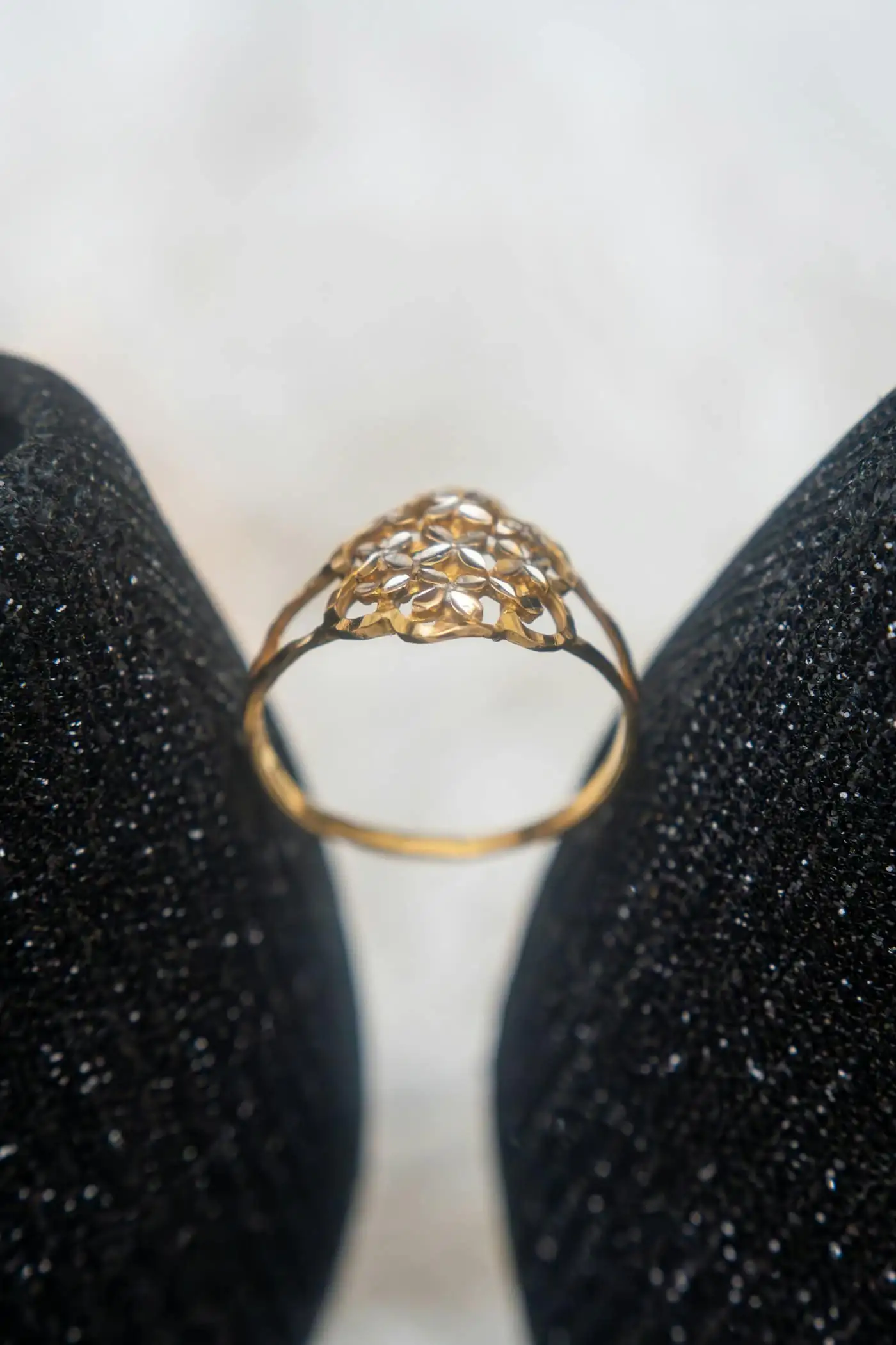 A gold filigree ring standing upright between a pair of black glittering shoes against a soft white background